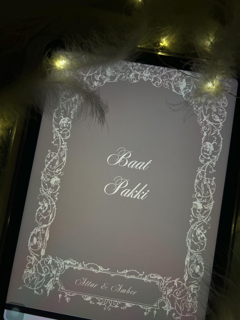 Puede incluir: Una tableta digital muestra una pantalla gris claro con un borde floral blanco y texto. El texto dice "Baat Pakki" y "Attar & Amber". La imagen est&aacute; enmarcada por un borde negro e iluminada por luces suaves y c&aacute;lidas y plumas blancas.