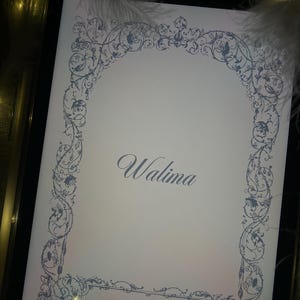 Puede incluir: Una tarjeta de invitaci&oacute;n blanca con un borde floral azul y la palabra "Walima" en escritura elegante. Los nombres "Attar & Amber" se muestran en la parte inferior. La tarjeta est&aacute; sobre una bandeja negra con plumas.