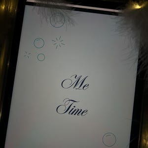 Puede incluir: Una tableta digital muestra las palabras "Me Time" en una elegante escritura. La pantalla es blanca con c&iacute;rculos y estrellas azules decorativas. La tableta est&aacute; rodeada de suaves plumas blancas y luces doradas c&aacute;lidas.