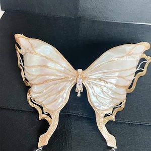 May include: A white and gold butterfly-shaped brooch with a crystal embellishment in the center. The wings are textured and have a pearlescent finish.