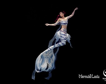 May include: A woman wearing a blue and white mermaid tail with a matching top poses underwater against a black background. The tail has a striped pattern and the top has a ruffled neckline. The woman has red hair and is wearing a red headband. The text "Mermaid Lucia" and a logo for "WaterLILY Mermaid" are visible in the bottom right corner of the image.
