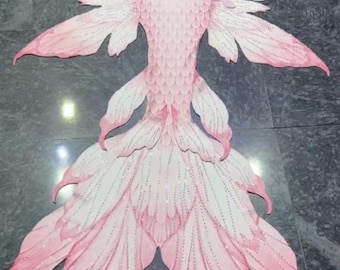 May include: A pink mermaid tail with a white and pink ombre effect. The tail is made of fabric and has a sparkly finish. The tail is laid out flat on a gray surface. The text "Mermaid Lucia" is visible in the bottom right corner of the image.