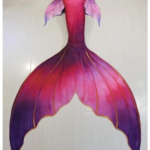 May include: A mermaid tail costume featuring a vibrant gradient of pink, magenta, and deep purple. The fabric displays a scale pattern on the upper section and translucent side fins. The large fluke has gold-colored trim and the text AAKI is visible.