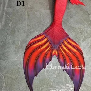 May include: Red, orange and purple mermaid tail with a fin that has a pointed tip. The tail is made of fabric and has a scaly texture. The tail is lying on a grey tiled floor. The text "D1" is visible in the top left corner of the image.