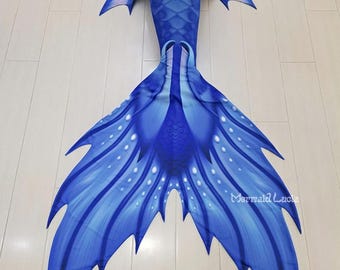 May include: A blue mermaid tail with a light blue and white pattern. The tail has a fin with a pointed tip and a scalloped edge. The tail is made of fabric and has a mermaid scale pattern. The tail is lying on a wooden floor. The text "S1" is visible in the upper left corner of the image.