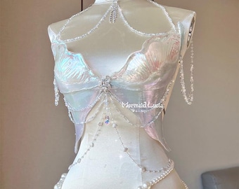 Könnte beinhalten: Ein weißer, schimmernder Meerjungfrauen-Kostüm-Oberteil mit Perlen- und Kristallketten-Detail. Das Oberteil hat ein muschelförmiges Design und der Text "Mermaid Luxia" ist auf der rechten Seite sichtbar.