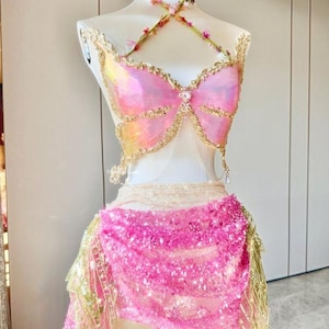 Könnte beinhalten: Ein zweiteiliges Set aus rosa und goldfarbenem Schmetterlingsoberteil und -rock. Das Oberteil hat schillernde Flügel und goldfarbene Verzierungen mit einem floralen Neckholder. Der Rock ist mit rosa Pailletten und goldenen Fransen bedeckt.
