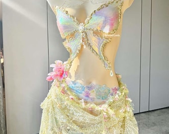 May include: A fantasy-themed costume displayed on a mannequin. The outfit features a butterfly-shaped bodice with iridescent wings and gold trim. A beaded necklace and a skirt with floral and lace details complete the ensemble. The color palette includes pastels and gold.