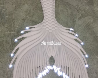 May include: White mermaid tail with light-up fins. The tail is made of fabric and has a scaly pattern. The fins have white lights that illuminate the tail.