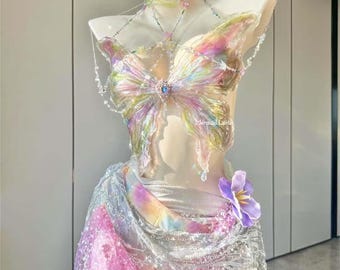 Puede incluir: Un top iridiscente con forma de mariposa con un escote halter de cuentas y una falda a juego con un estampado tie-dye y lentejuelas. La falda tiene un adorno de flor morada.