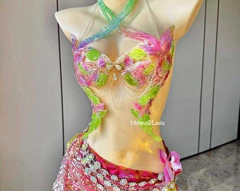 Puede incluir: Un colorido disfraz hecho a mano con un diseño floral. La parte superior presenta un cuello halter y está adornada con flores rosas, verdes y moradas. La falda es un diseño en capas con telas rosas y amarillas, adornada con lentejuelas y cuentas. El texto "Mermaid Lucia" es visible.