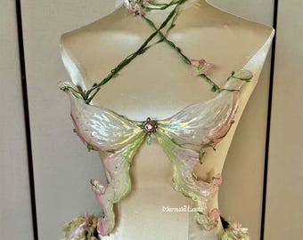 Puede incluir: Un disfraz de hada blanco y verde con un corpiño en forma de mariposa y un collar de vid verde con flores rosas. El disfraz está en un maniquí blanco. El texto "Mermaid Loxa" es visible en la parte inferior de la imagen.
