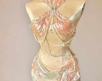 Puede incluir: Un disfraz de sirena exhibido en un maniquí. El disfraz presenta una falda con lentejuelas y una parte superior con detalles iridiscentes de color coral. Delicados hilos de perlas y una joya central añaden detalles. El texto "Mermaid Lucia" es visible.