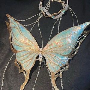 May include: A pair of iridescent butterfly wings with gold accents and crystal chains. The wings are attached to a gold metal headband with a crystal design. The wings are a light blue and green color with a shimmery finish. The headband is a delicate gold metal with a crystal design. The chains are made of clear crystals and are attached to the wings and headband. The wings are a beautiful and delicate piece of jewelry that would be perfect for a costume or special occasion.