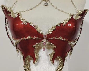 Puede incluir: Un corpiño rojo y dorado con forma de mariposa con detalles intrincados. El corpiño presenta un acabado rojo brillante, detalles de filigrana dorada y un delicado collar de cadena adornado con cristales y un colgante central. Incluye adornos de cristal colgantes.