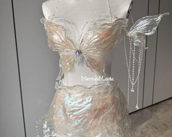 May include: A white iridescent costume with a butterfly design. The costume features a top, skirt, and wings. The top and wings are made of a sheer fabric with a butterfly design. The skirt is made of a more opaque fabric with a flowing design. The costume is decorated with iridescent beads and crystals.