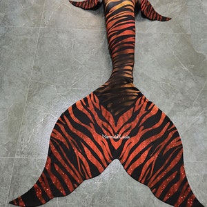 May include: A mermaid tail with a tiger stripe design in shades of orange and black. The tail features a split fin design and is decorated with small white dots. The text "Mermaid Lucia" is visible on the fin.