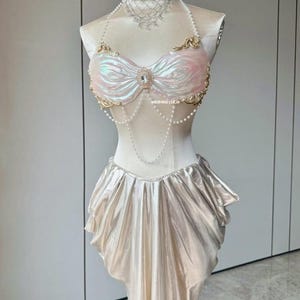 May include: A mermaid-themed costume with a pearlescent top and skirt. The top features a bow design with gold trim and a central jewel. A pearl necklace and choker complete the look. The skirt has a draped, flowing design.