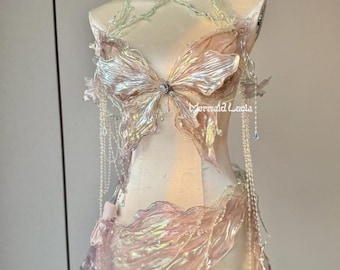 May include: A white and iridescent butterfly-shaped bra with a matching skirt. The bra and skirt are made of a sheer, flowing fabric with a delicate, ethereal look. The bra has a crystal embellishment in the center. The skirt is layered and has a flowing, mermaid-like design. The text "Mermaid Lucia" is visible on the skirt.