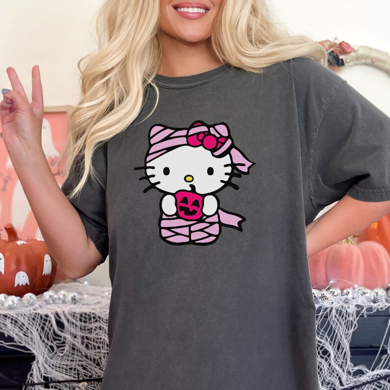 May include: A dark grey t-shirt featuring a graphic of Hello Kitty dressed as a mummy holding a jack-o'-lantern.