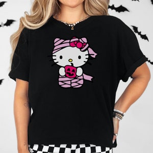 May include: Black t-shirt featuring a pink Hello Kitty graphic. Hello Kitty is dressed as a mummy and holding a pumpkin.