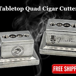 May include: Silver tabletop cigar cutter with four cutting blades. The cutter has a United States Marine Corps logo engraved on the front. The cutter is on a black background with smoke.