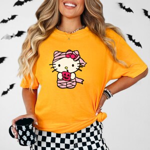 May include: A yellow t-shirt featuring a cartoon image of Hello Kitty dressed as a mummy holding a jack-o-lantern.