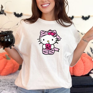 May include: White t-shirt featuring a pink Hello Kitty graphic. Hello Kitty is dressed as a mummy and holding a jack-o-lantern.
