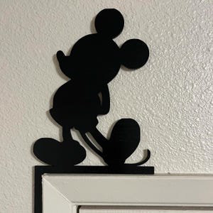 Puede incluir: Una silueta negra del personaje de dibujos animados Mickey Mouse, diseñada como pieza decorativa para esquinas. La figura aparece en una pose clásica, de pie sobre una base plana situada en la esquina superior de un marco de puerta blanco.