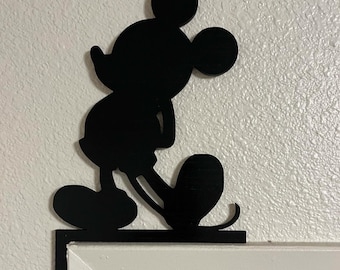 May include: A black silhouette of the cartoon character Mickey Mouse, designed as a corner accent piece. The figure is depicted in a classic pose, standing on a flat base that is positioned along the top corner of a white door frame.