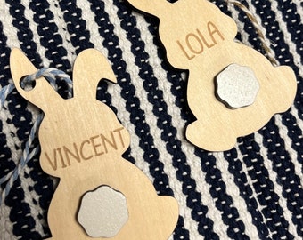 Op de afbeelding: Twee houten bunny-vormige ornamenten met de namen "Lola" en "Vincent" erop gegraveerd. Elk ornament heeft een witte, ronde, vilt-achtige versiering aan de achterkant.