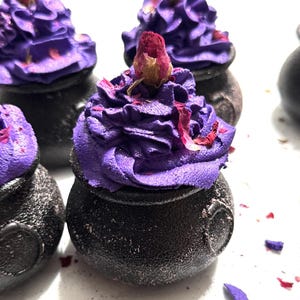 Könnte beinhalten: Nahaufnahme von mehreren schwarzen, kesselartigen Badesalzen, die mit lila Zuckerguss und einer getrockneten Rosenknospe bedeckt sind. Die Badesalze sind mit lila Streuseln und Rosenblättern verziert. Der Hintergrund ist weiß.