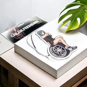 May include: A white car air freshener with a cartoon image of a woman sitting on a tire. The woman has green hair and is wearing a black dress and white sneakers. The air freshener is on a white box.