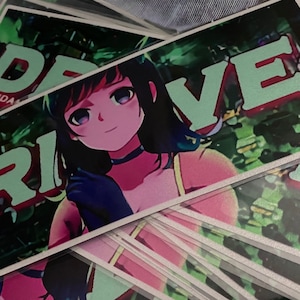 May include: A stack of stickers featuring a colorful anime-style illustration of a girl with long black hair and a green and white text overlay that reads "DRIVE".