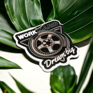 May include: A white sticker with a black and white tire design and the text "Work Hard Dream Big".