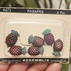 May include: A package of six miniature pineapple decorations. The pineapples are brown with blue-green tops, and one is purple. The package is labeled "PINEAPPLE" and "international ASSEMBLIX corporation."