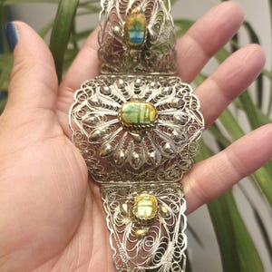 May include: A silver filigree bracelet with three sections, each featuring a colorful scarab beetle design. The bracelet has an ornate, openwork pattern and is held in a hand.