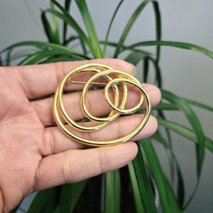 May include: A gold-toned brooch featuring three interlocking circular rings of varying sizes. The polished metal surface reflects light, creating a smooth, continuous loop design. The brooch is constructed with a pin mechanism on the back side.