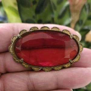 May include: An antique brooch featuring a large, faceted, oval-shaped red glass stone. The stone is set in a gold-toned metal frame with a decorative scalloped edge. The brooch is a vintage piece, likely from the early 20th century.