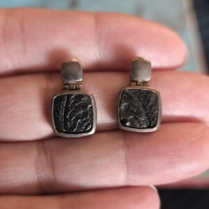 May include: A pair of silver-toned earrings with a square, textured black center. The earrings have a rectangular top and a square bottom with a leaf-like design. The earrings are shown against a blurred background.