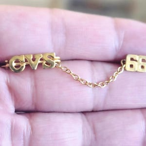 May include: A gold-toned pin with the letters "CVS" connected by a delicate chain to a small square with the number "66". The pin is likely vintage or a commemorative piece, with a classic design.