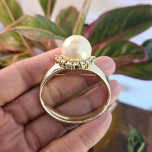 May include: A gold-toned ring brooch featuring a large, cream-colored pearl at the center. The pearl is surrounded by a halo of small, clear crystals. The brooch has a simple, elegant design, suitable for adding a touch of sophistication to any outfit.