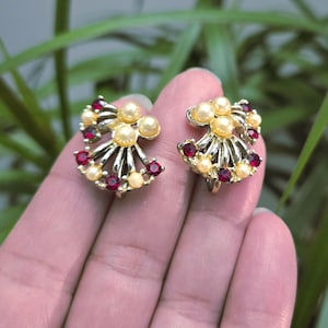 May include: A pair of vintage clip-on earrings. Each earring features a gold-toned fan design with faux pearls and small, round, red gemstones. The earrings are held in a hand against a blurred green background.
