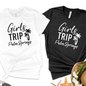 以下が含まれることがあります： 「Girls Trip Palm Springs」の文字とヤシの木のグラフィックがプリントされたTシャツ2枚。1枚は白、もう1枚は濃いグレーです。