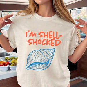 May include: Cream-colored t-shirt with the text "I'M SHELL-SHOCKED" in orange and a blue seashell graphic. The shirt is a casual, short-sleeved design, perfect for beach or summer wear.