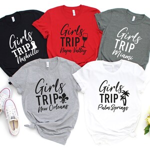 以下が含まれることがあります： 「Girls Trip」の文字と各目的地がプリントされたTシャツが5枚。目的地はナッシュビル、ナパバレー、マイアミ、ニューオーリンズ、パームスプリングスです。