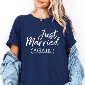 May include: Navy blue t-shirt with the white text "Just Married (AGAIN)". The shirt has rolled-up sleeves and is worn with a denim jacket. The text is in a cursive font.