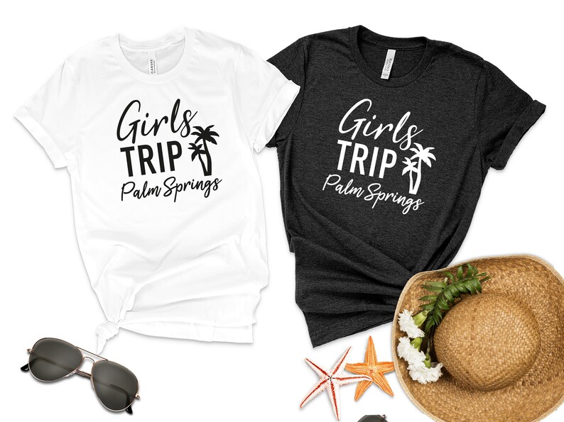 以下が含まれることがあります： 「Girls Trip Palm Springs」の文字とヤシの木のグラフィックがプリントされたTシャツ2枚。1枚は白、もう1枚は濃いグレーです。