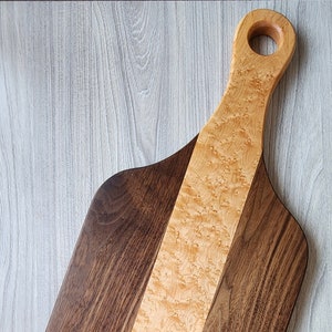 May include: A wooden chopping board with a light brown centre stripe and dark brown sides. The board has a handle with a hole in it. The board is made of walnut and maple wood. "Generation Home Decor" is printed on the bottom right corner of the board.
