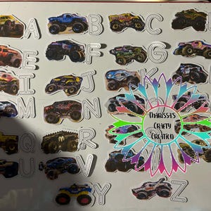 May include: A set of alphabet stickers, each featuring a different monster truck design. The trucks are in various colours, including yellow, blue, and red. The image also includes a decorative flower with the text "Darissa's Crafty Creation".